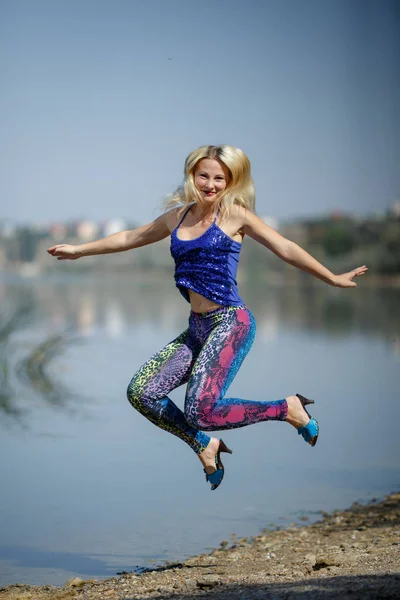 young smiling girl in colored leggings and blue top on heels jumps on a river shore