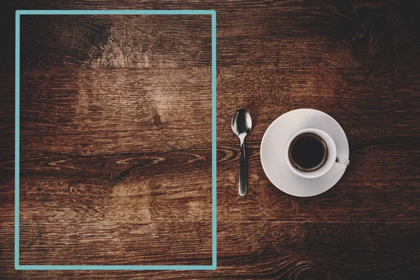 Vista superior de la taza blanca con café negro en un platillo blanco y cucharadita sobre fondo de madera marrón oscuro con marco azul a la izquierda —  Fotos de Stock