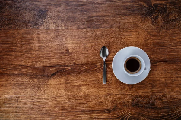 Draufsicht auf weiße Tasse mit schwarzem Kaffee auf weißer Untertasse und Teelöffel auf dunkelbraunem Holzhintergrund — Stockfoto