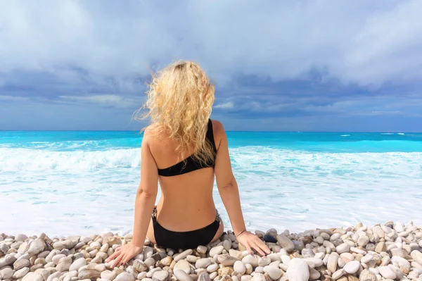 Schöne blonde Mädchen in einem schwarzen Badeanzug sitzt an einem leeren karibischen Strand — Stockfoto