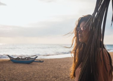 Sahilin arka planında güneş gözlüklü bir kız. Teknesi ve okyanusu var.