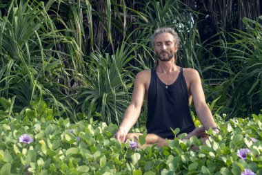Kafasında rasta olan erkek bir yogi tropik yeşillik ve çiçeklere karşı lotus pozisyonunda oturur..