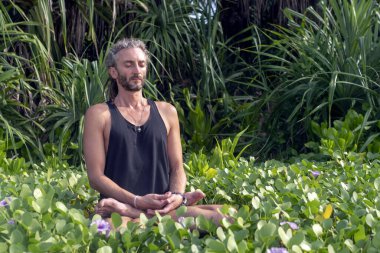 Kafasında rasta olan erkek bir yogi tropik yeşillik ve çiçeklere karşı lotus pozisyonunda oturur..