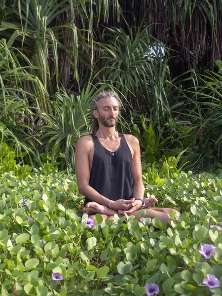 Kafasında rasta olan erkek bir yogi tropik yeşillik ve çiçeklere karşı lotus pozisyonunda oturur..