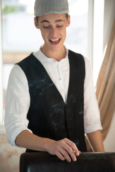 Молодой человек в кепке и жилете курит у окна
