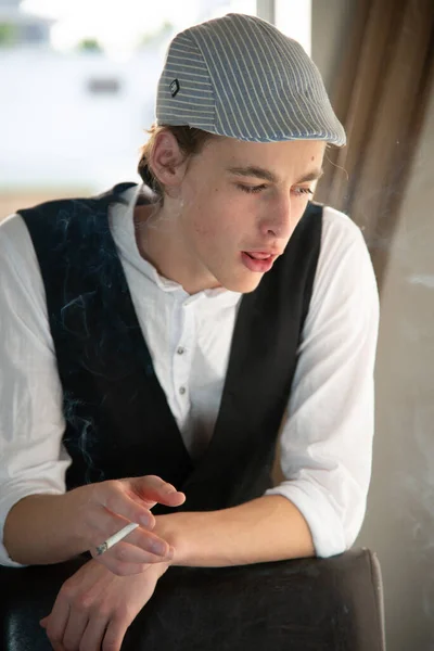 Młody mężczyzna w czapce i kamizelce pali przy oknie — Zdjęcie stockowe