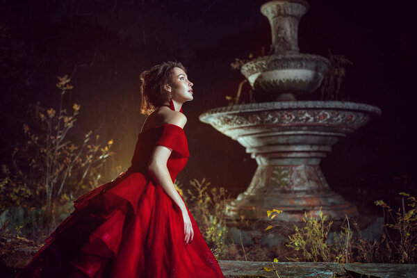 Beautiful lady dressed in red ball gown staying near the fountain