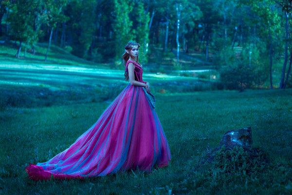 Beautiful young elf woman dressed in long purple dress walking through the summer forest