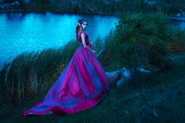 Beautiful young elf woman dressed in long purple dress walking through the summer forest