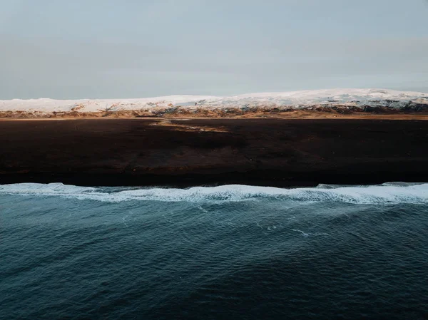 The coast of Iceland with a black sand beach and some mountains in the background, photographed from a drone