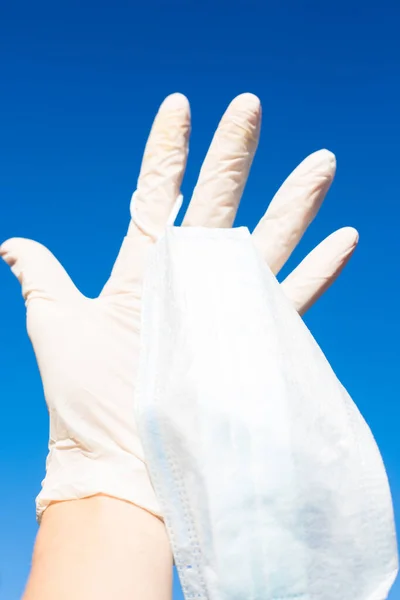 Human hand in white medical glove with open palm on intense blue background. Medical mask hang on forefinger