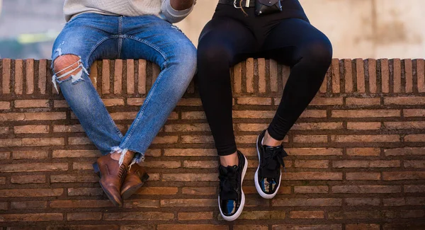 Pár sedí na cihlové římské zdi nedefinovaný. Muži v modrých džínách a hnědých botách, žena v černých kamaších a teniskách, oranžová stěna jako lavička — Stock fotografie