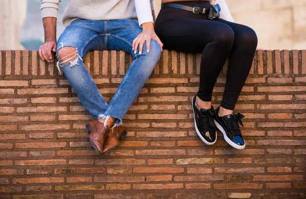 Tuğla duvarda oturan çift tanımlanmamış. Kot pantolonlu ve kahverengi çizmeli erkekler, siyah taytlı ve spor ayakkabılı kadınlar, sıralı turuncu duvarlar.