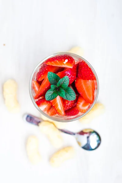 Üstünde kırmızı çilek parçaları ve taze nane yaprağı olan bir bardak. Beyaz ahşap arka planda yuvarlak bir tatlıya üst manzara.