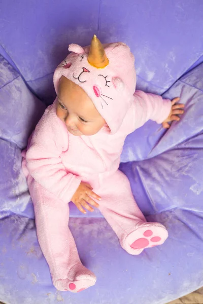 Pembe tek boynuzlu at kostümlü bebek. Çocuk açık mor renkli şişme sandalyede tek başına oturuyor ve yan tarafa bakıyor. İtalyan karnavalı. Bebeklerin doğum günü partisi