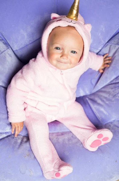 Pembe tek boynuzlu at kostümlü bebek. Çocuk açık mor renkli şişme sandalyede tek başına oturuyor ve doğrudan kameraya bakıyor. İtalyan karnavalı. Bebeklerin doğum günü partisi