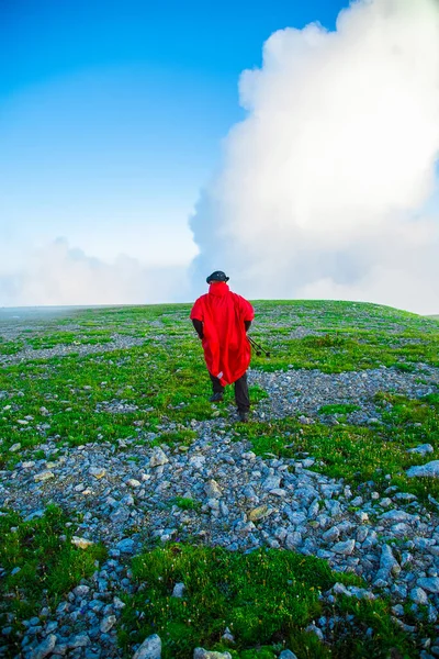 Alp Vadisi 'ndeki parlak kırmızı ceketli dağcı. Dağın tepesinde. Bulutların önündeki zümrüt tarlasında kırmızılı adam. Yazın Apennines 'te yürüyüş. İzolasyon konsepti. Sıra dışı turizm 