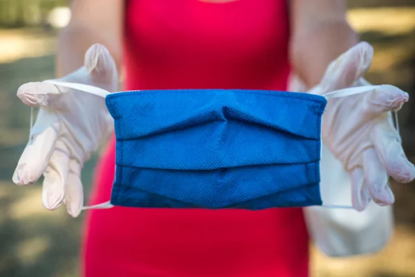 Pembe elbiseli kadın, el yapımı mavi cerrahi koruyucu bir maske gösteriyor. Günlük hayatta tıbbi maske ve beyaz eldivenler. Karantina gerçeğinden sonraki yaz günü. Maske takmak zorundalar. Parkta yaz günü 