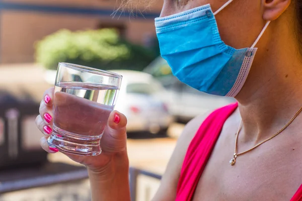 Mavi tıbbi maskeli kız elinde bir bardak saf su tutuyor. Pandemik dönemde içki içmek. Covid 'den sonra halka açık yerlerde koruyucu maske takmak. İtalyan barında yaz günü 