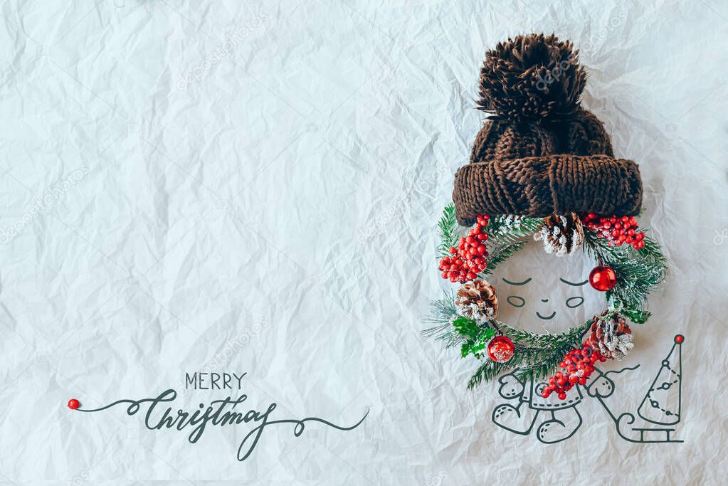 Christmas concept. Cute baby made of wreath of evergreen spruce, knitted hat and illustration on a white background. Minimal winter vacation idea. Flat lay top view composition.