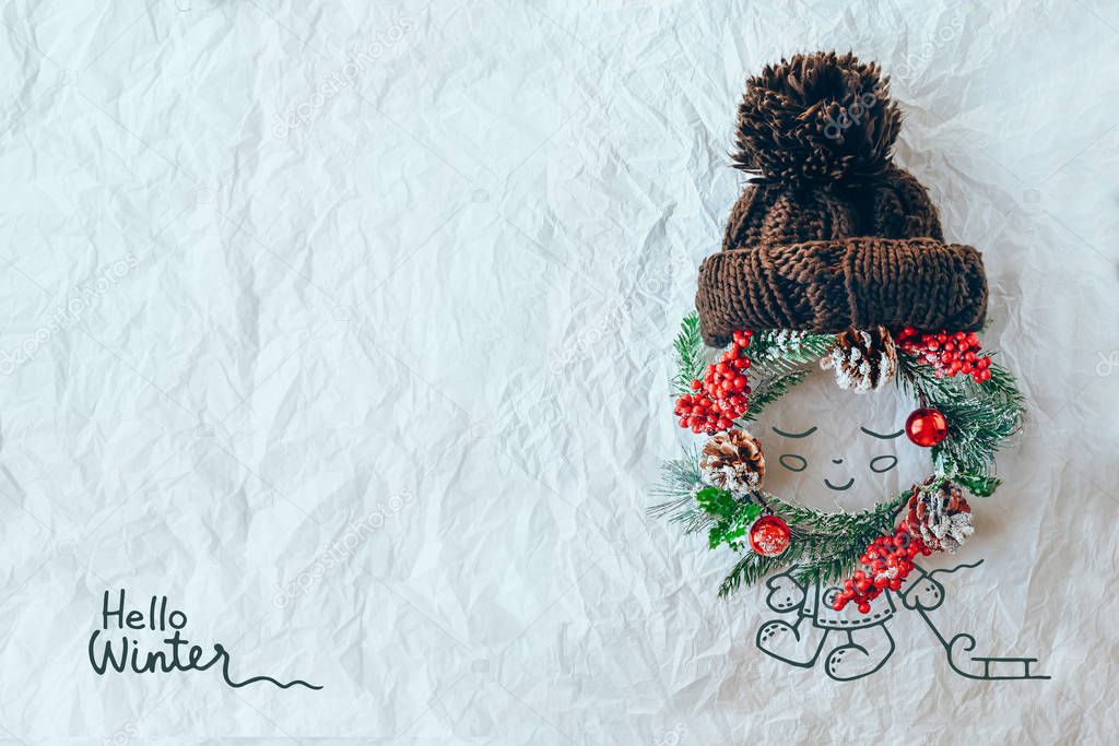 Christmas concept. Cute baby made of wreath of evergreen spruce, knitted hat and illustration on a white background. Minimal winter vacation idea. Flat lay top view composition.