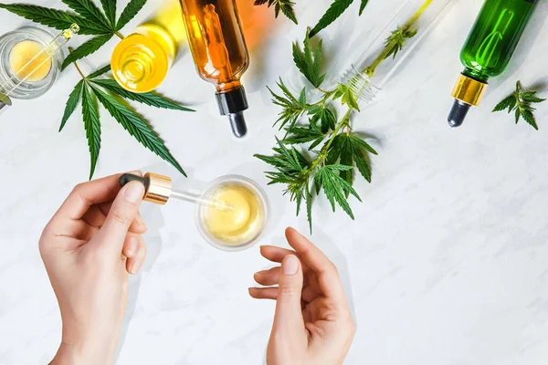 Varias botellas de vidrio con aceite de CBD, tintura y hojas de cáñamo sobre un fondo de mármol. Manos femeninas sosteniendo una pipeta con aceite cosmético CBD — Foto de Stock