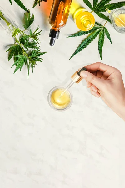 Піпетт з косметичною олією CBD в жіночих руках на столі з різними пляшками з конопляною олією і листям коноплі, марихуана. — стокове фото