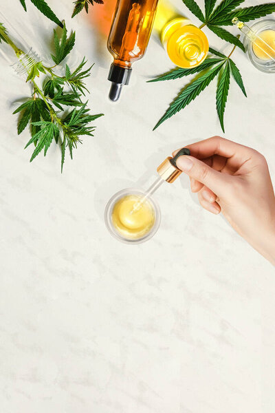 Трубка с CBD косметическим маслом в женских руках на фоне стола с различными бутылками с марихуаной и коноплей, марихуана

