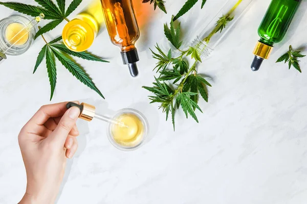 Pipeta com óleo cosmético CBD em mãos femininas em um fundo de mesa com cosméticos, creme com folhas de cannabis e cânhamo, maconha — Fotografia de Stock