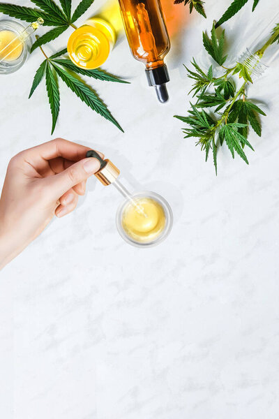 Различные стеклянные бутылки с CBD масла, настойки и конопли листья на мраморном фоне. Руки женщины держат пипетку с косметическим маслом CBD
