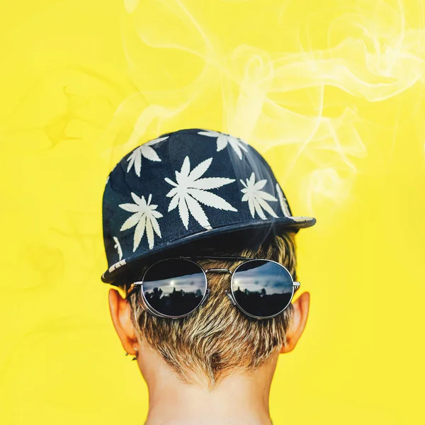 Menina em óculos de sol e boné de beisebol com folhas de maconha fuma em um fundo de parede amarelo. Fumo de cânhamo — Fotografia de Stock