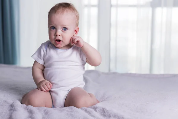 Un bebé sorprendido con la boca abierta. Rascarse detrás de la oreja niño pequeño sentado en la cama frente a la ventana —  Fotos de Stock