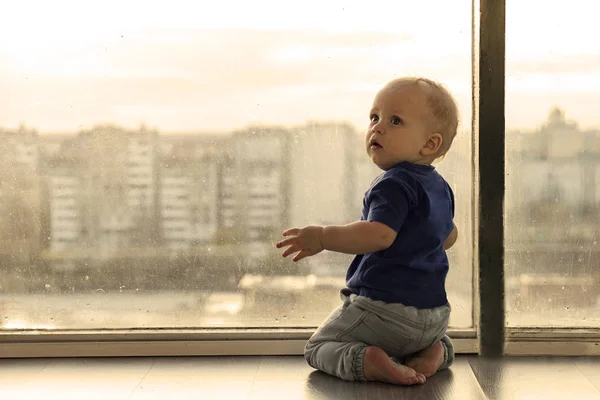 Ładny chłopczyk patrząc przez okno do deszczowa wielkiego miasta. Niemowlę chłopiec czeka na rodziców. Toddler dziecko siedzi przez okno i wygląda zdenerwowany. — Zdjęcie stockowe