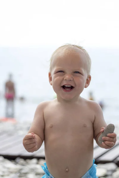 Joyeux petit garçon souriant sur la plage. Joli bambin riant sur un bord de mer regardant la caméra. Fond flou — Photo