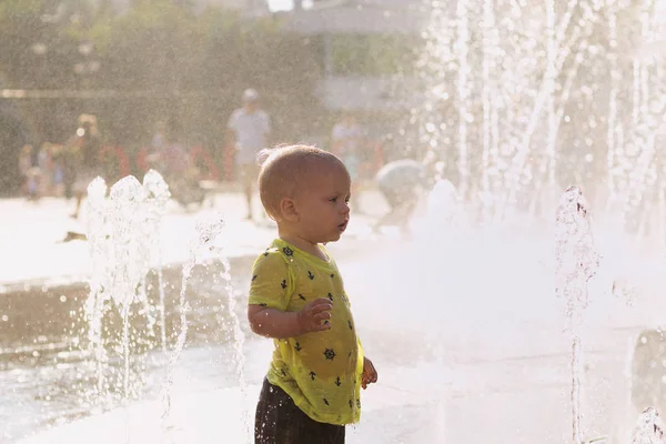 Милый малыш в мокрой одежде стоит в центре городского фонтана. Капли воды, распыляемые в воздухе . — стоковое фото