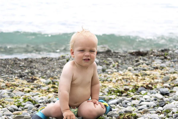 Ładna przez chłopiec siedzi na kamienistą plażę. Zabawne dziecko nad brzegiem morza, patrząc na kamery — Zdjęcie stockowe