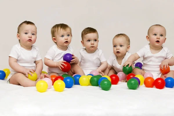 Riga di bambini caucasici seduti fianco a fianco guardando dall'altra parte isolati su sfondo grigio. Cinque simpatici bambini che giocano con le palle colorfull — Foto Stock