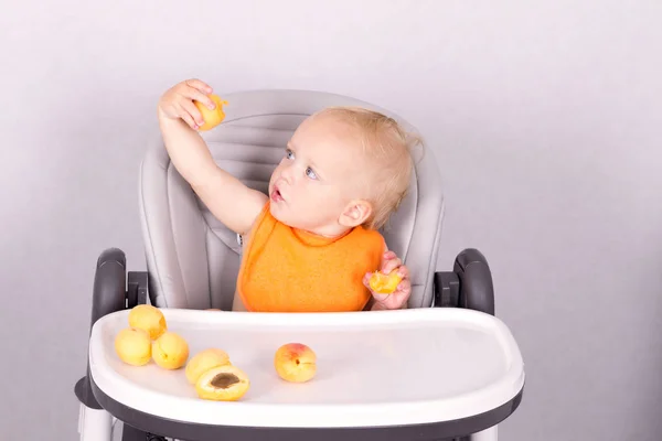Очаровательный малыш ест абрикос в детском кресле на сером фоне — стоковое фото