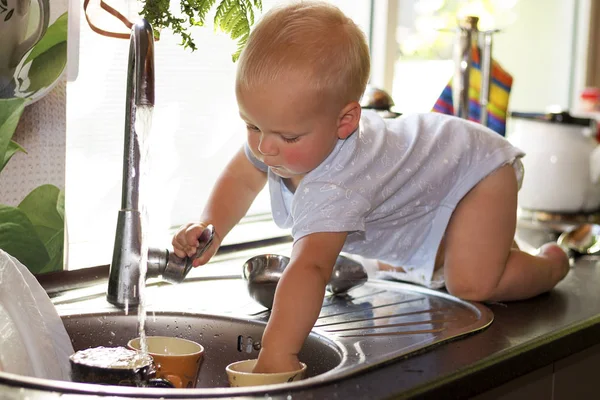 Милий маленький хлопчик миє посуд на домашній кухні. Чарівний хлопчик грає з водяним краном — стокове фото