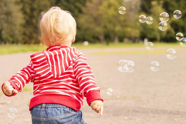 Милый малыш, бегающий среди мыльных пузырей в парке. Вид сзади — стоковое фото