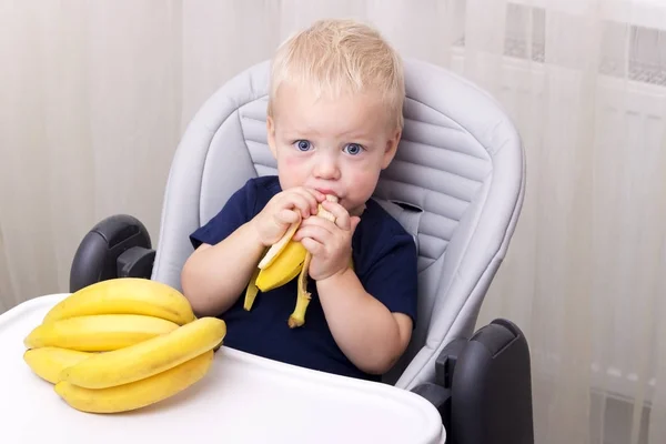 Милый годовалый малыш ест банан и сидит в детском кресле . — стоковое фото