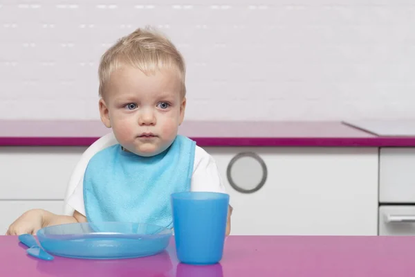 Portrait Of thoughtful adorable little kid in a blue bib In High Chair in the modern kitchen ready to eat. Copy space — Stock Photo, Image