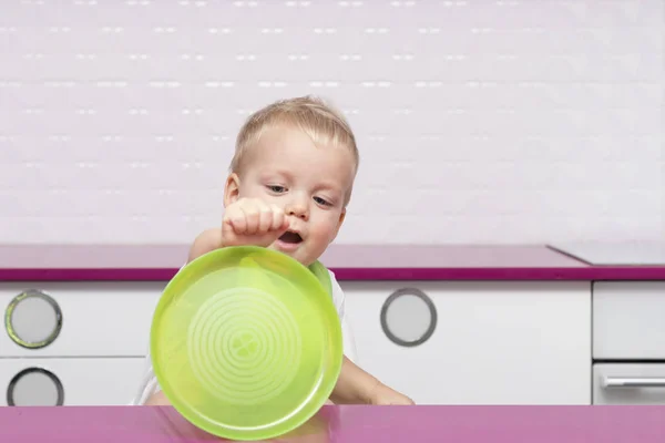 Милый мальчик играет с зеленой пластиковой пластиной на современной белой кухне. Забавный малыш показывает свою пустую тарелку . — стоковое фото