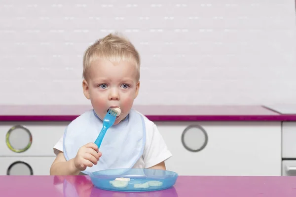 Милый малыш в синем нагруднике ест банан на современной кухне — стоковое фото