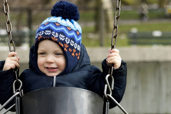 Счастливого улыбающегося малыша на качелях на детской площадке. Весна или осень — стоковое фото
