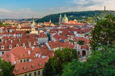 Prag, Çek Cumhuriyeti - 20 Temmuz 2019: Kızıl çatılar, kiliseli şehir manzarası ve kulesi olan uzak Petrin tepesi. Bulutlu mavi akşam gökyüzü. Önplanda yeşil ağaçlar.