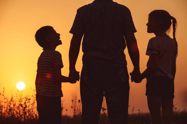 Baba ve çocuklar gün batımında parkta dikiliyorlar. İnsanlar sahada eğleniyor. Arkadaş canlısı aile kavramı ve yaz tatili.