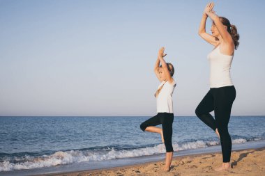 Anne ve kızı gündüz vakti plajda yoga egzersizleri yapıyor. İnsanlar dışarıda eğleniyor. Yaz tatili ve arkadaş canlısı aile kavramı.