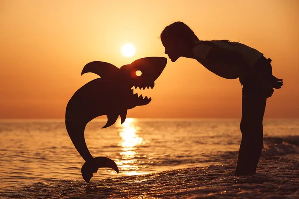 夕日の時間にビーチに立って1人の幸せな女の子 彼女は段ボールのサメと遊ぶ 子供は屋外で楽しんでいる 夏休みの概念 — ストック写真