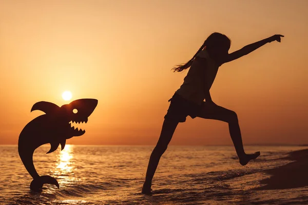 夕日の時間にビーチを走っている幸せな女の子が一人います 彼女は段ボールのサメと遊ぶ 子供は屋外で楽しんでいる 夏休みの概念 — ストック写真